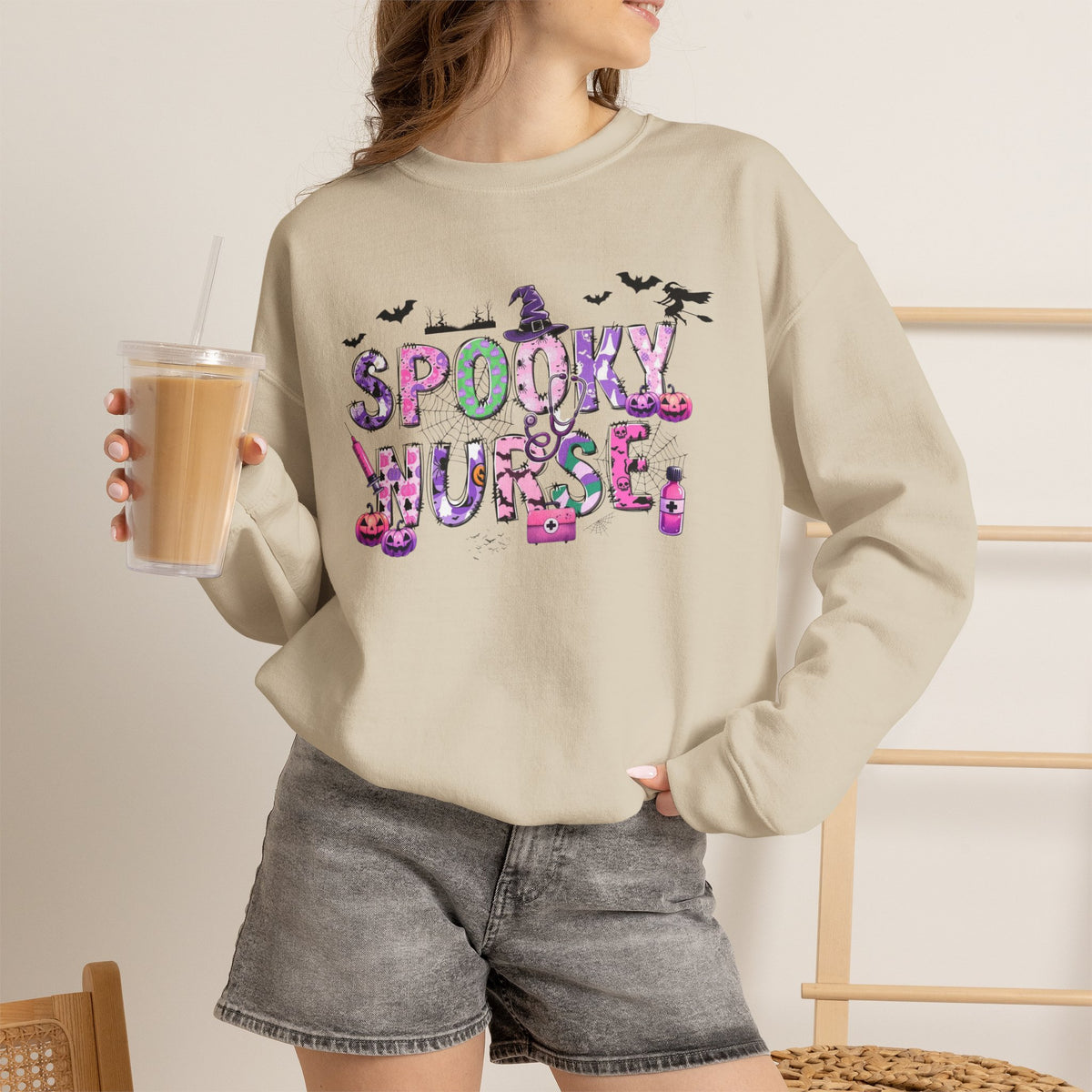 A woman wearing a beige crewneck sweatshirt with a colorful 'Spooky Nurse' graphic, paired with gray denim shorts and holding an iced coffee in a bright indoor setting.