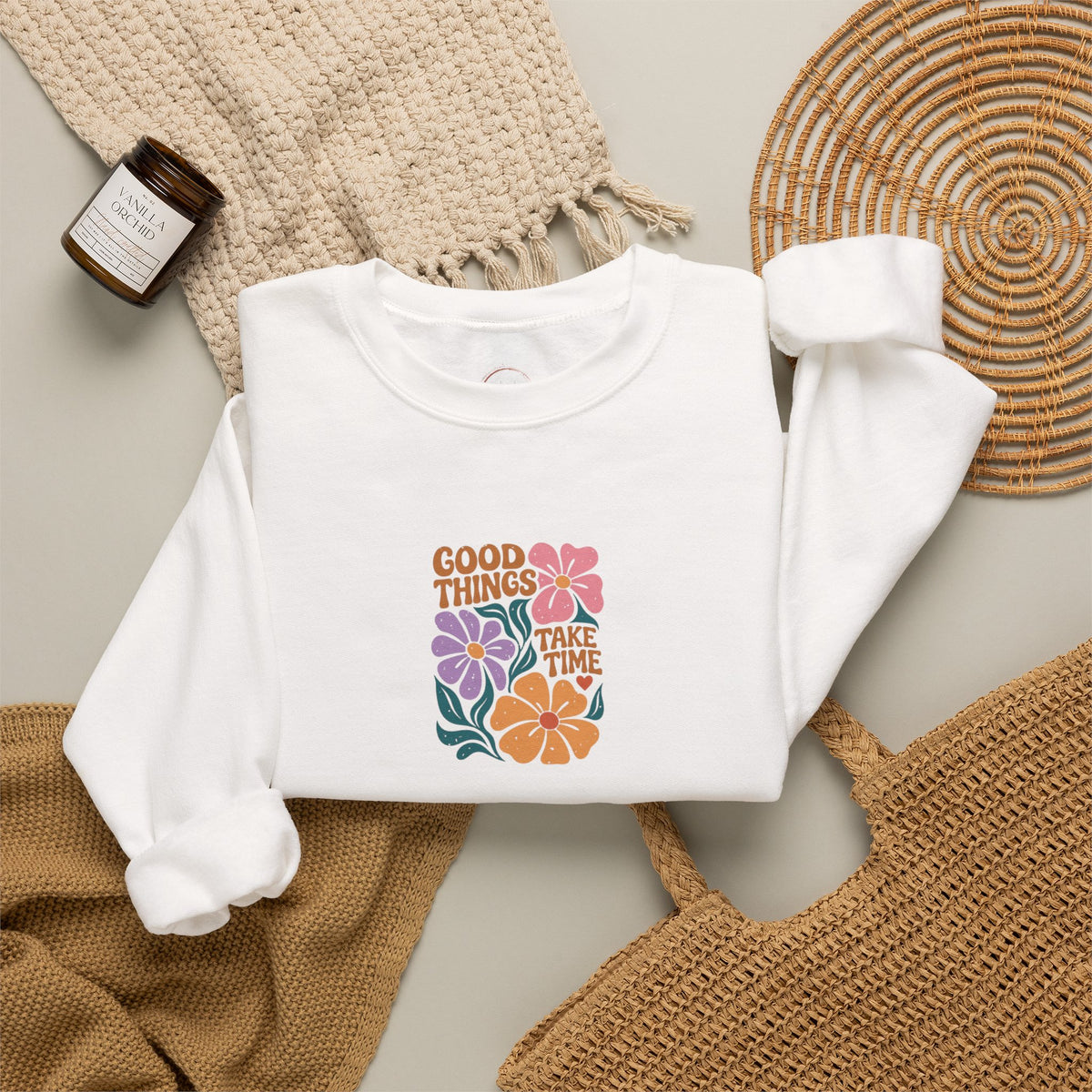 A folded white long-sleeve sweatshirt with a colorful retro graphic reading 'GOOD THINGS TAKE TIME' surrounded by flowers, styled in a flat lay with a woven blanket, a small candle jar, and a wicker tray on a light neutral background.