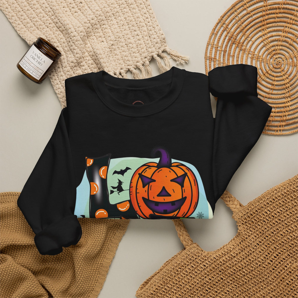 A black long-sleeve crewneck sweatshirt with a colorful Halloween graphic print featuring a jack-o'-lantern, bats, and pumpkins, styled in a flat lay with a beige knit blanket, a woven placemat, and a small candle.