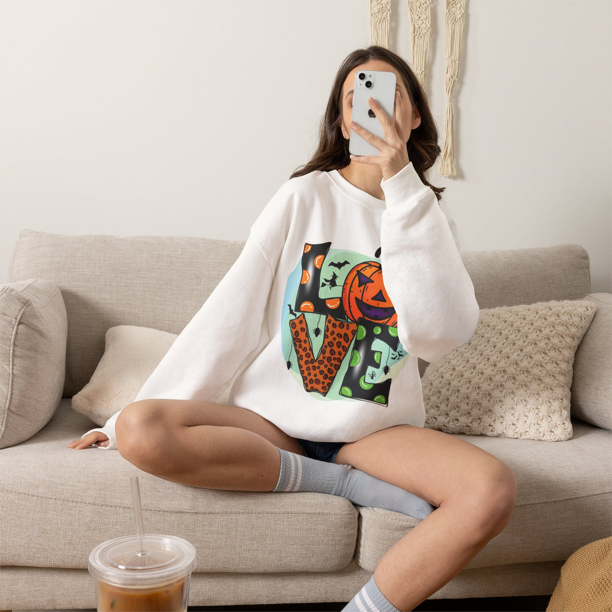 A woman sitting on a beige couch takes a mirror selfie while wearing a white crewneck sweatshirt with a colorful graphic print of the word 'LOVE' in a stylized, geometric, retro font. The print features orange, green, black, and red details.