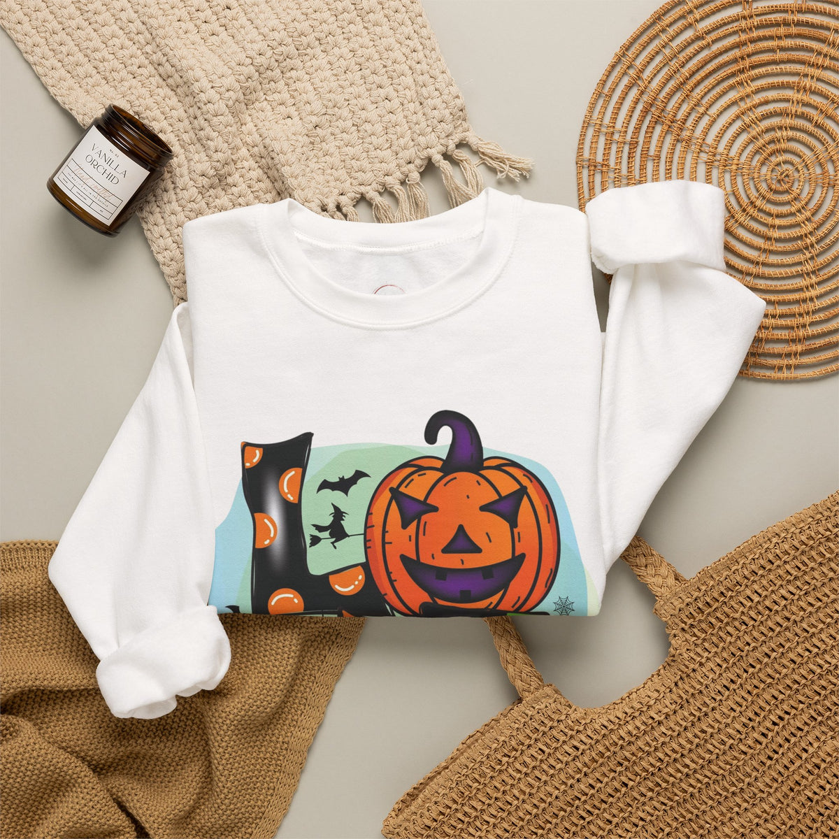 A white long-sleeve crewneck sweatshirt with a colorful Halloween graphic print featuring a jack-o'-lantern, a witch's hat, bats, and pumpkins, styled in a flat lay with a knit blanket, woven placemat, and a candle.