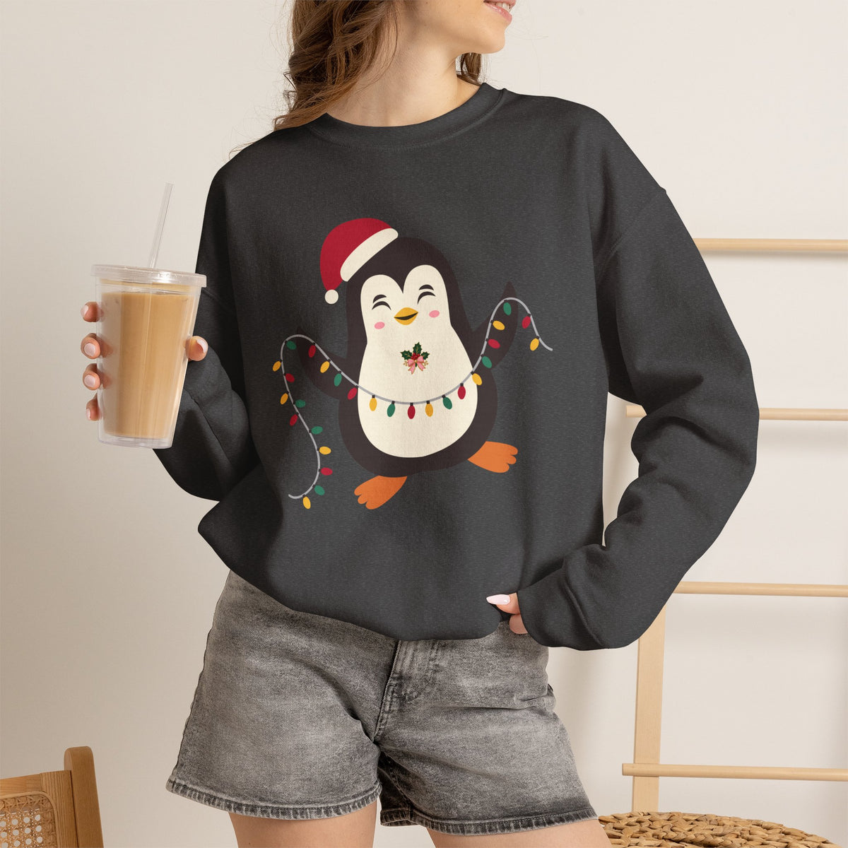 A woman wearing a dark charcoal gray crewneck sweatshirt with a festive cartoon penguin graphic, holding a iced coffee in a bright indoor setting.