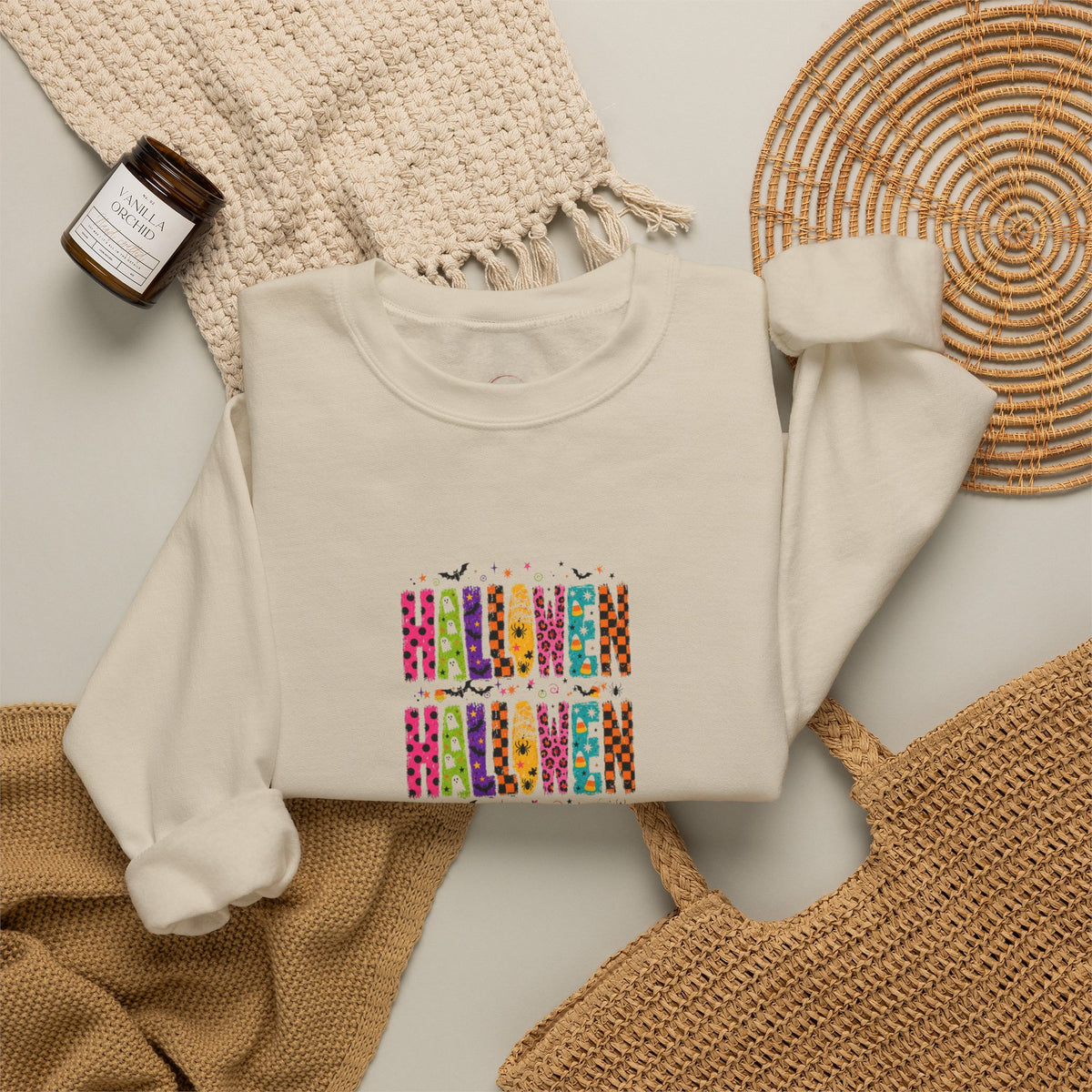 An off-white crewneck sweatshirt with a colorful 'HAPPY HALLOWEEN' graphic print, styled in a flat lay with a beige knit blanket, a woven placemat, a small candle, and a tan knit bag on a light gray background.