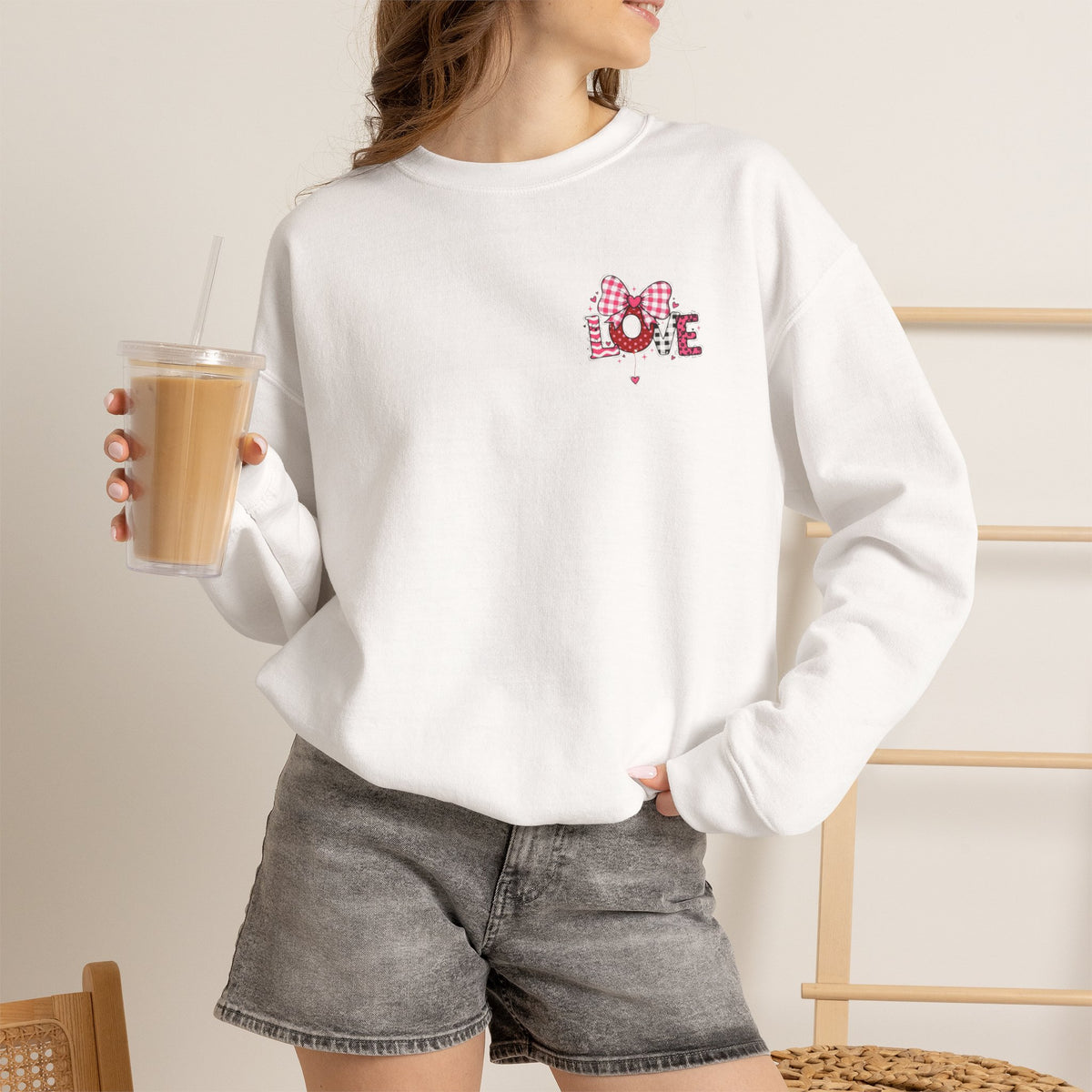 A woman wearing a white crewneck sweatshirt with a small graphic print on the chest, paired with gray denim shorts and holding an iced coffee in a cafe setting.