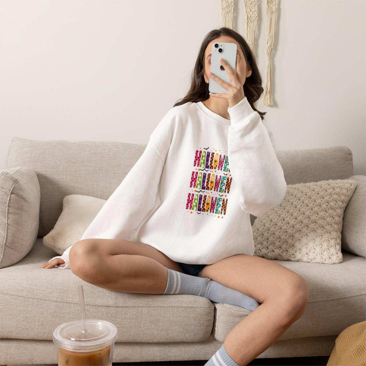 A woman sitting on a beige couch takes a mirror selfie while wearing a white crewneck sweatshirt with a colorful graphic print on the front. She is wearing gray striped socks and holding a phone, with an iced coffee in the foreground.