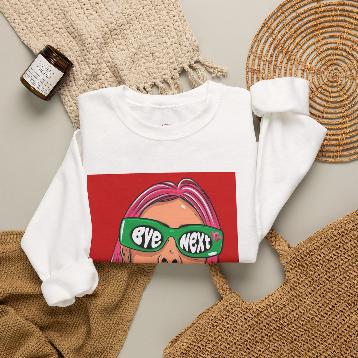 A white crewneck sweatshirt laid flat on a neutral background, featuring a graphic print of a cartoon face with pink hair and green sunglasses that read 'eye next'. The sweatshirt is styled with a beige knit blanket, a woven placemat, a small candle jar, and a tan woven bag.