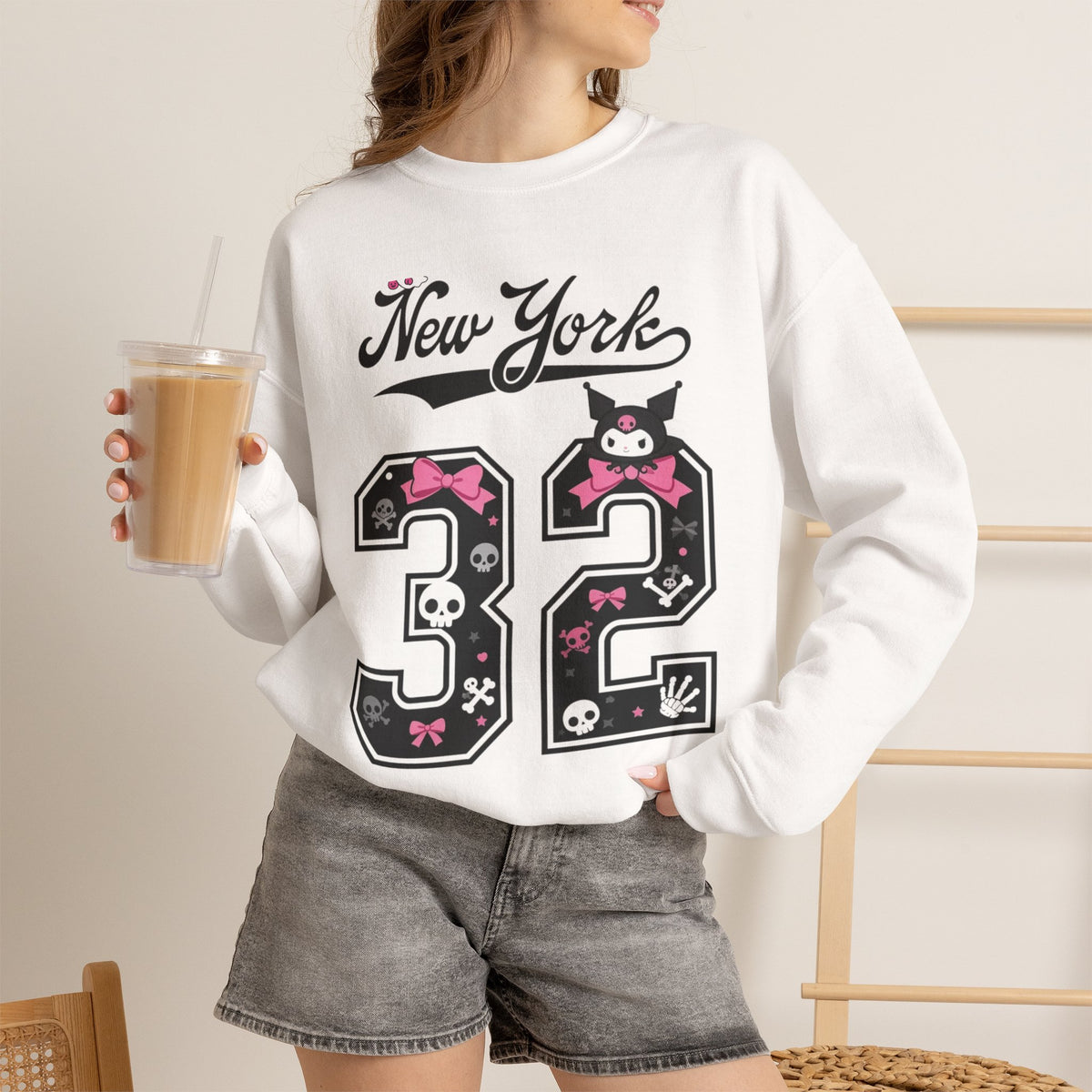 A woman wearing a white crewneck sweatshirt with a black and pink 'New York 32' graphic featuring cartoon character details, paired with gray denim shorts and holding an iced coffee.