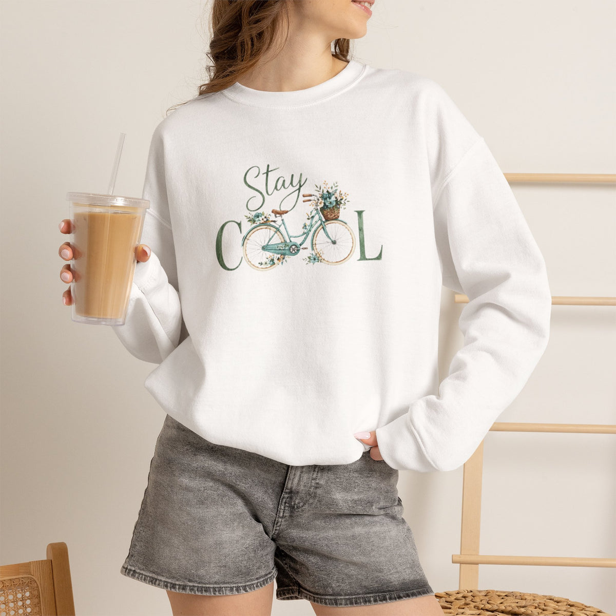 A woman wearing a white crewneck sweatshirt with a 'Stay COOL' bicycle graphic, paired with gray denim shorts and holding an iced coffee.