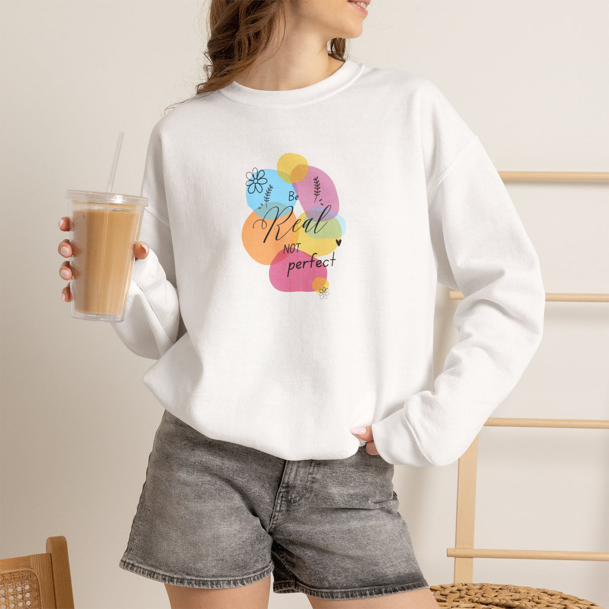 A woman wearing a white crewneck sweatshirt with a colorful graphic that reads 'Rude, not perfect' in a pastel balloon-style design. She is holding an iced coffee and wearing gray denim shorts in a brightly lit indoor setting.