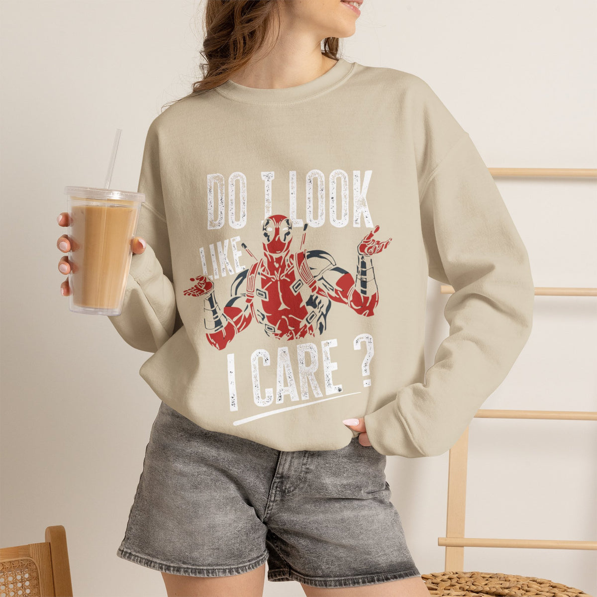 A woman wearing a beige crewneck sweatshirt with a Deadpool graphic and the text 'DO I LOOK LIKE I CARE?' while holding an iced coffee. She is wearing gray denim shorts in a brightly lit indoor setting.