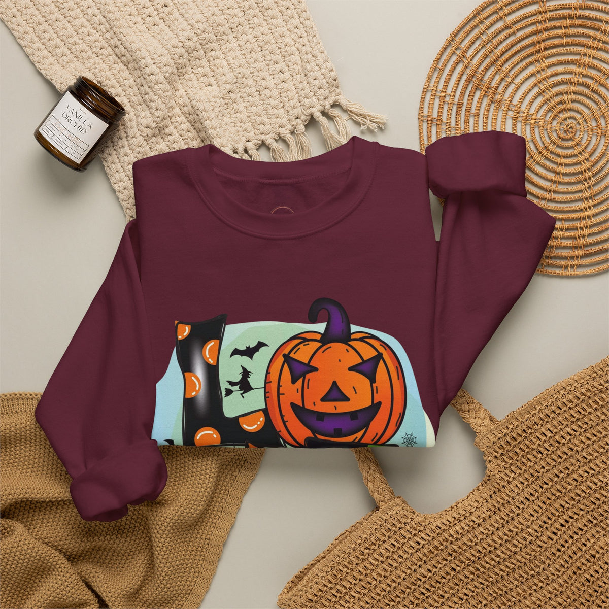 A maroon crewneck sweatshirt with a colorful Halloween graphic of a jack-o'-lantern, bats, and pumpkins, styled in a flat lay with a beige knit blanket, a woven placemat, a straw bag, and a candle.