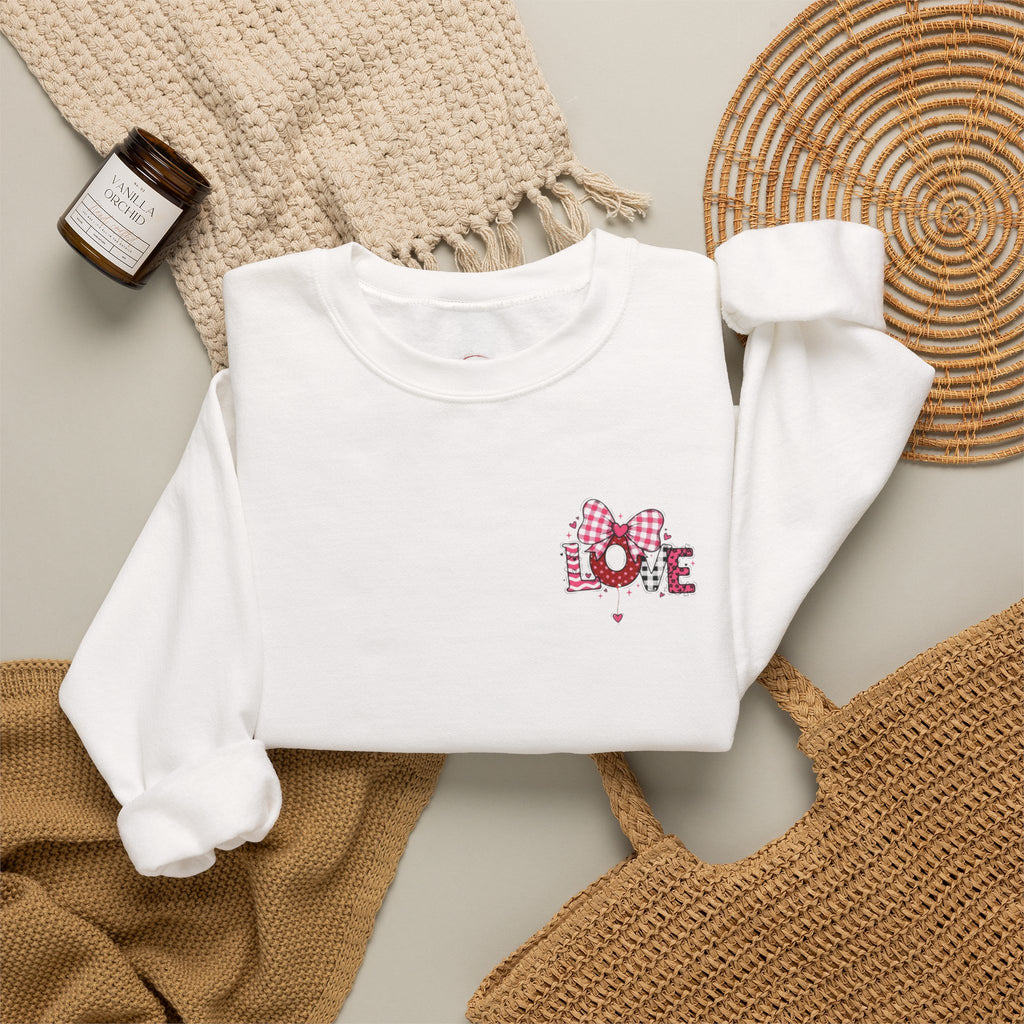 A white long-sleeve crewneck sweatshirt laid flat on a light gray surface, styled with beige knit textiles, a woven round mat, and a small amber candle. The sweatshirt has a small chest graphic reading LOVE in pink gingham and glittery pink lettering with a pink bow.