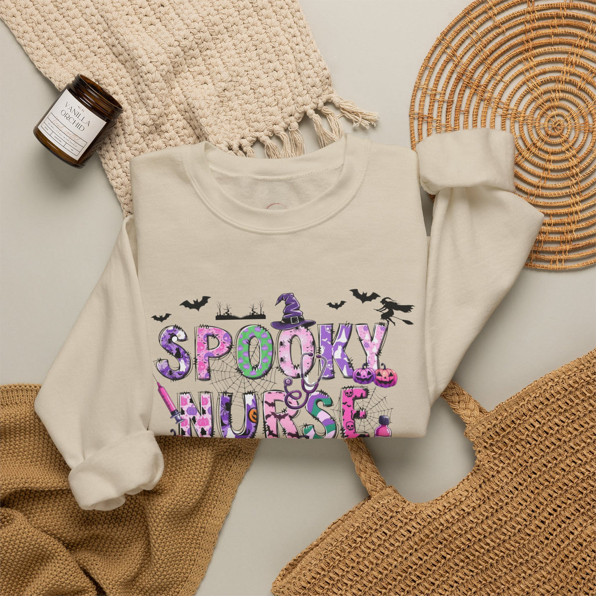 An off-white crewneck sweatshirt with a colorful 'Spooky Nurse' graphic, styled in a flat lay with a woven placemat, a knit blanket, a straw hat, and a candle.