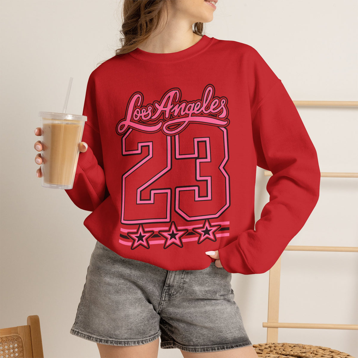 A woman wearing a red crewneck sweatshirt with a colorful floral wreath graphic and the text 'Genesis 50:22' printed on the front. She is holding an iced coffee and wearing gray denim shorts.