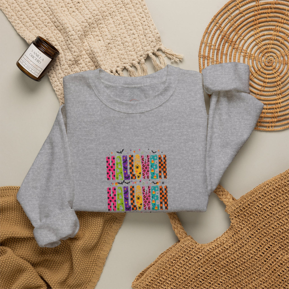 A folded heather gray sweatshirt with a colorful graphic print of stacked patterned plates and small bats, styled in a flat lay with a woven blanket, a candle jar, a wicker tray, and a tan knit bag on a light neutral background.