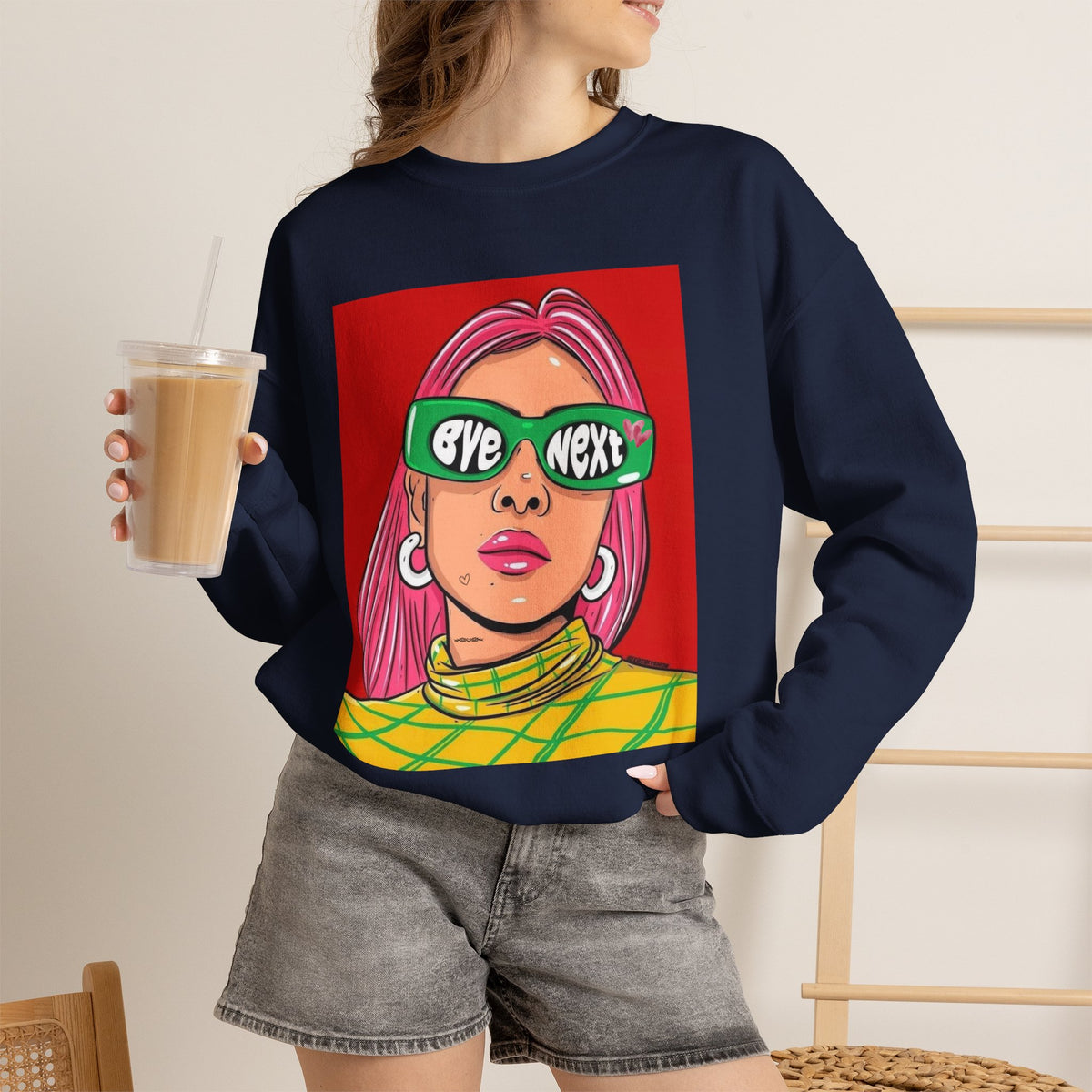 A woman wearing a navy blue crewneck sweatshirt with a colorful graphic print of a woman with pink hair and green sunglasses that read 'EYE NEXT'. She is holding an iced coffee and wearing gray denim shorts.