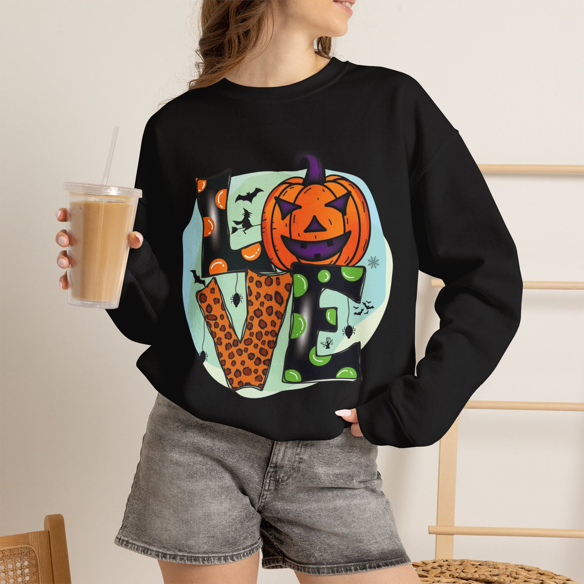 A woman wearing a black crewneck sweatshirt with a colorful Halloween-themed graphic spelling 'LOVE' featuring a jack-o'-lantern and bats, paired with gray denim shorts and holding an iced coffee.