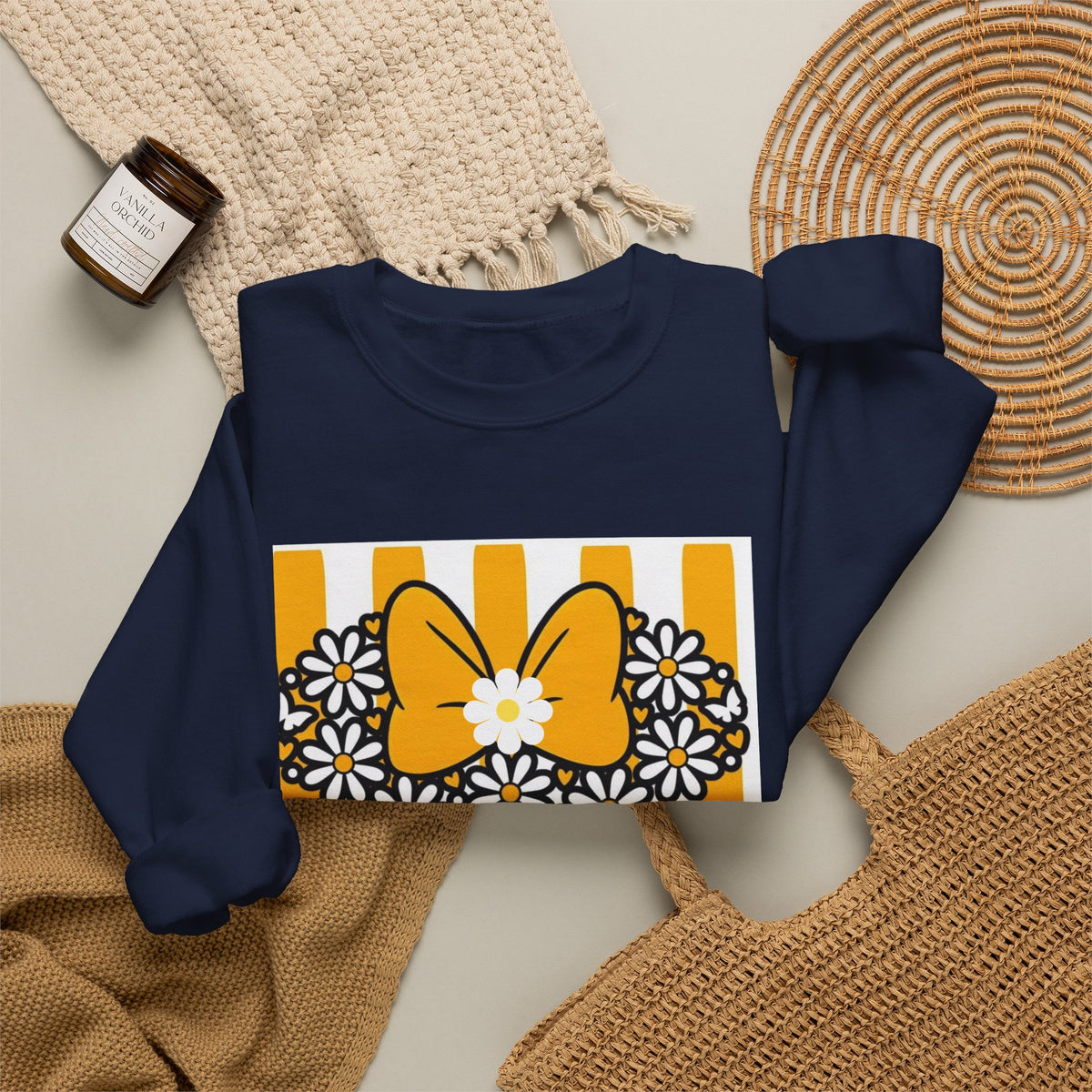 A navy blue sweatshirt laid flat on a light beige surface, styled with a cream knit blanket, a woven placemat, a tan tote bag, and a small candle. The sweatshirt features a large graphic print on the front with a yellow bow, white daisies, and yellow-and-white vertical stripes.