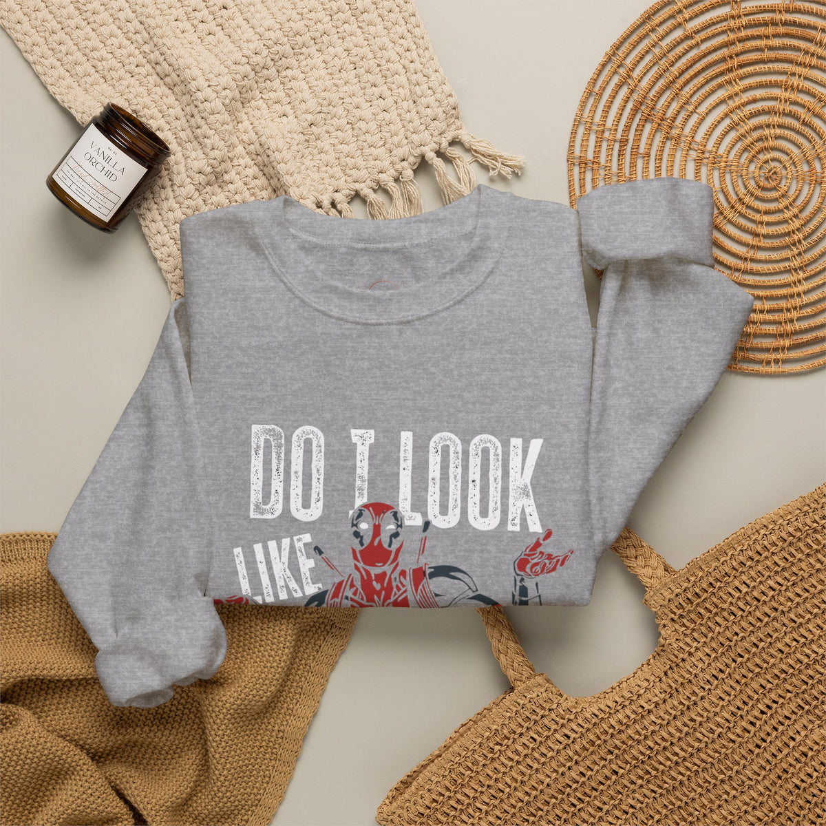 A heather gray crewneck sweatshirt with a graphic print of Deadpool and the text 'DO I LOOK LIKE' in distressed white letters, styled in a flat lay with a woven blanket, a candle, and a wicker tray.