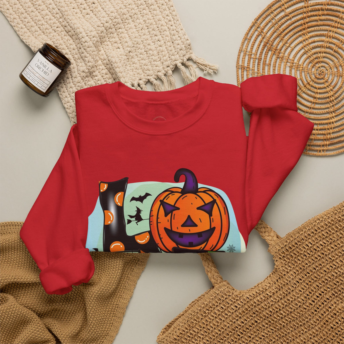 A red long-sleeve crewneck sweatshirt laid flat on a light beige surface, featuring a colorful Halloween graphic with a jack-o'-lantern, witch hat, bats, and a ghost. The sweatshirt is styled with a cream knit blanket, a woven placemat, a tan knit fabric, and a small candle jar.