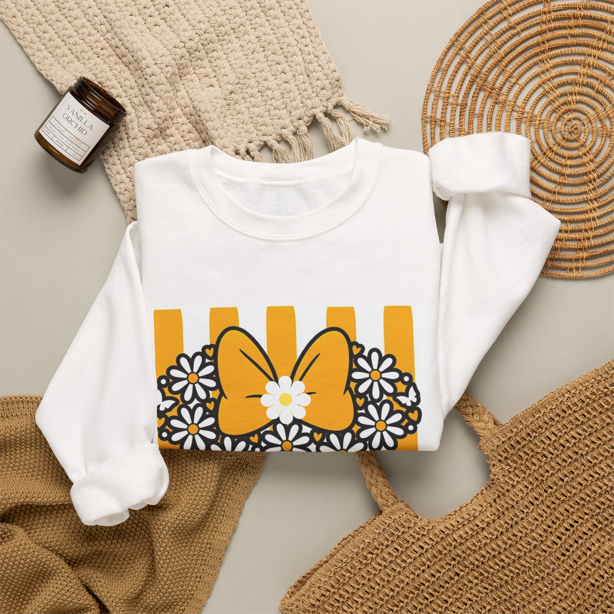 A white long-sleeve sweatshirt laid flat on a neutral background with styled props, featuring a graphic print of a yellow bow, white daisies, and yellow vertical stripes.