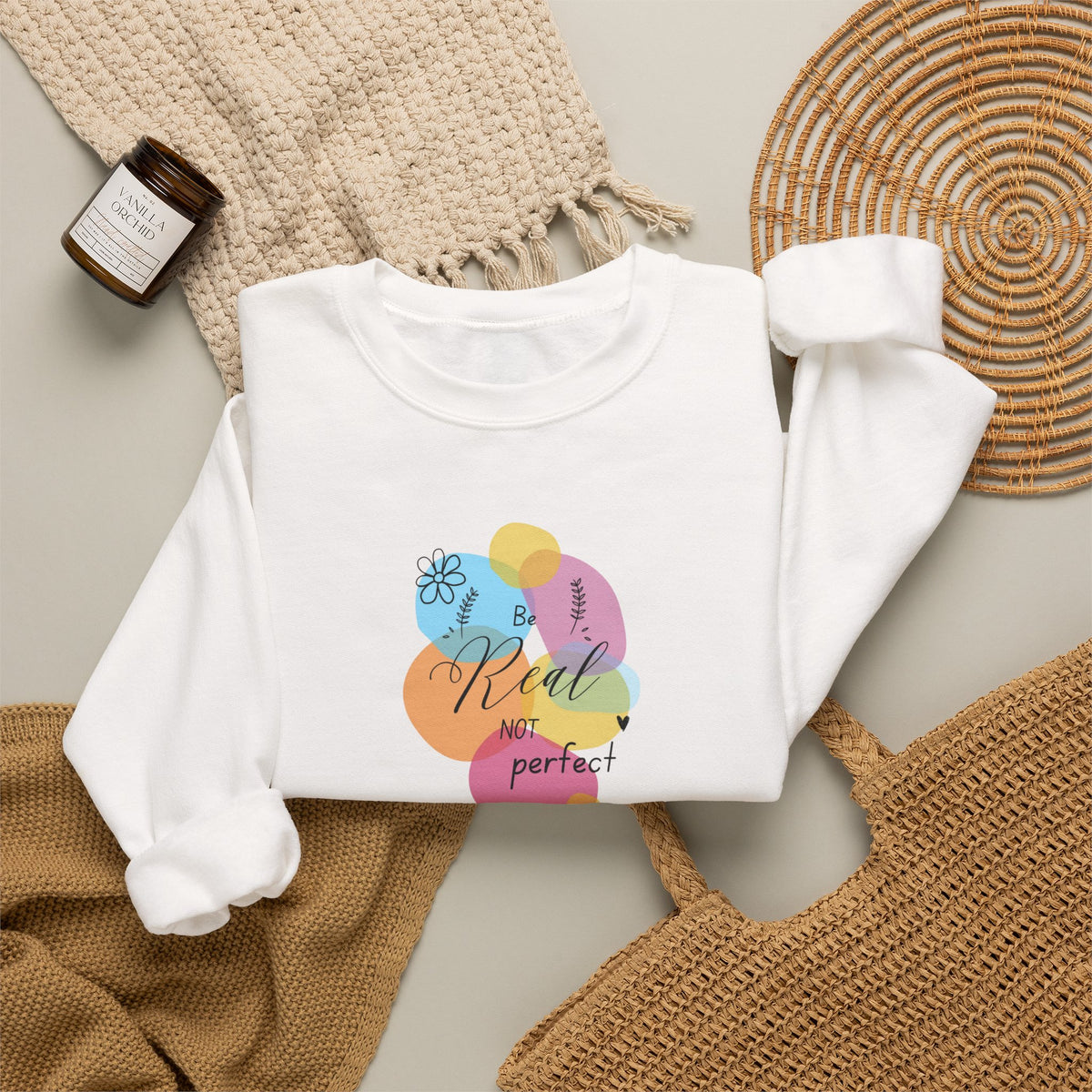 A folded white long-sleeve sweatshirt with a colorful graphic print reading 'Be Real NOT perfect' over pastel abstract shapes, styled in a flat lay with a woven blanket, a small candle jar, and a round woven mat on a light neutral background.