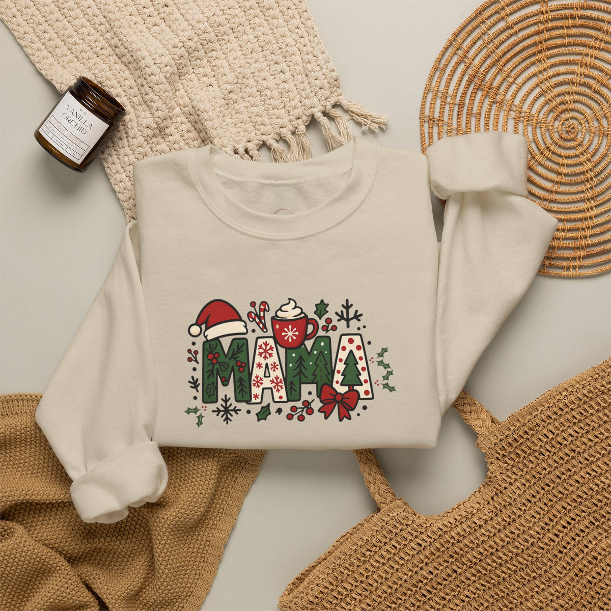 An off-white crewneck sweatshirt with a festive 'MAMA' graphic featuring Christmas-themed illustrations, styled in a flat-lay arrangement with a knit blanket, a woven mat, and a small candle.