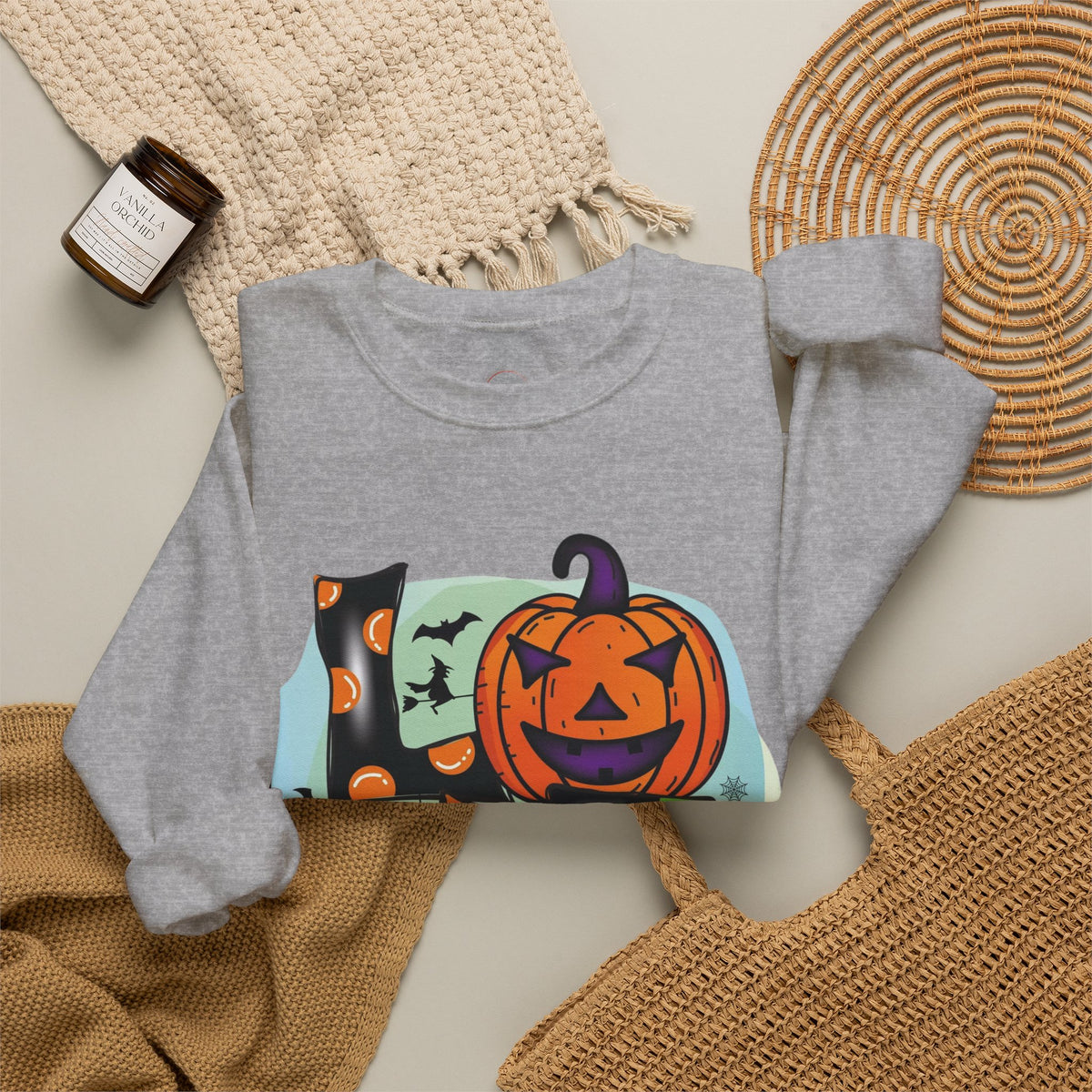 A heather gray crewneck sweatshirt with a colorful Halloween graphic print featuring a jack-o'-lantern, a witch's hat, bats, and a full moon, styled in a flat lay with a woven blanket, a small candle, and a wicker placemat.