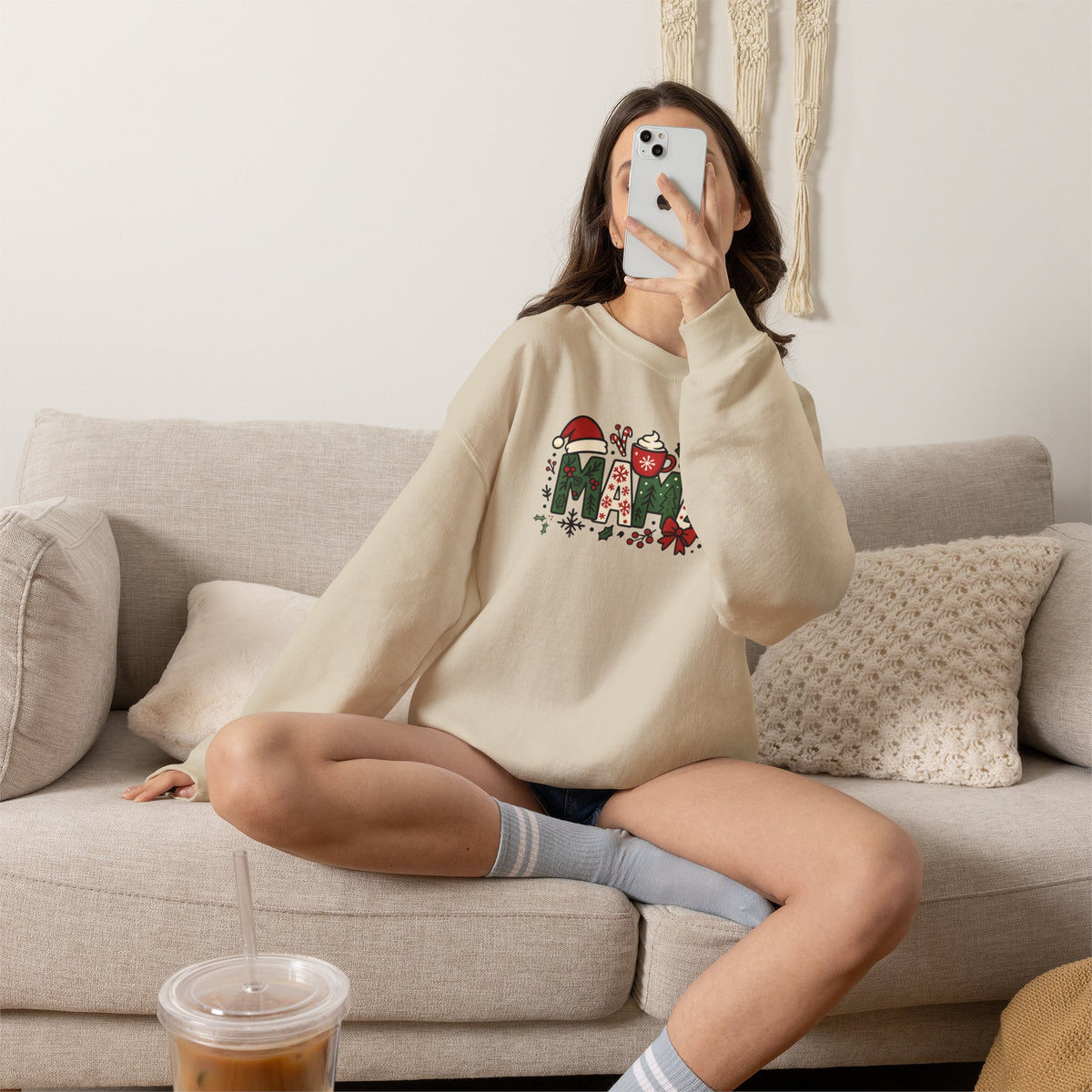A woman takes a mirror selfie while sitting cross-legged on a light beige couch. She is wearing a cream-colored crewneck sweatshirt with a colorful graphic of three corgis on the front, paired with gray striped socks and dark shorts.
