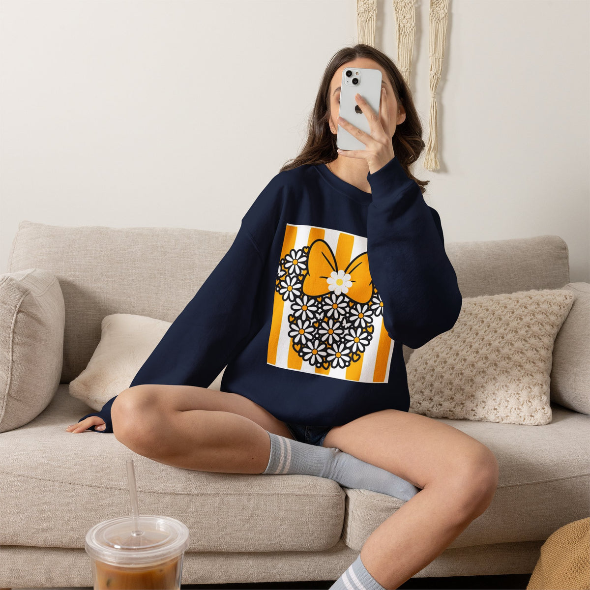 A woman takes a mirror selfie while sitting on a beige couch, wearing a navy blue sweatshirt with a graphic print of a gift box and a phone. An iced coffee is visible in the foreground.