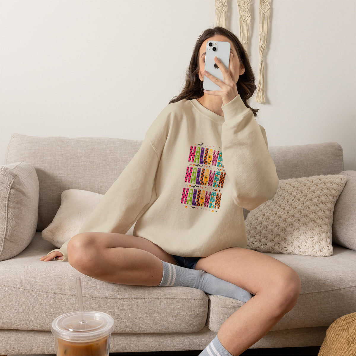 A woman sitting on a beige couch takes a mirror selfie while wearing a beige sweatshirt with a pink graphic print that reads 'DON'T BE PINK FOOL YOU' and 'I FAIL IT IN THE AMI'.