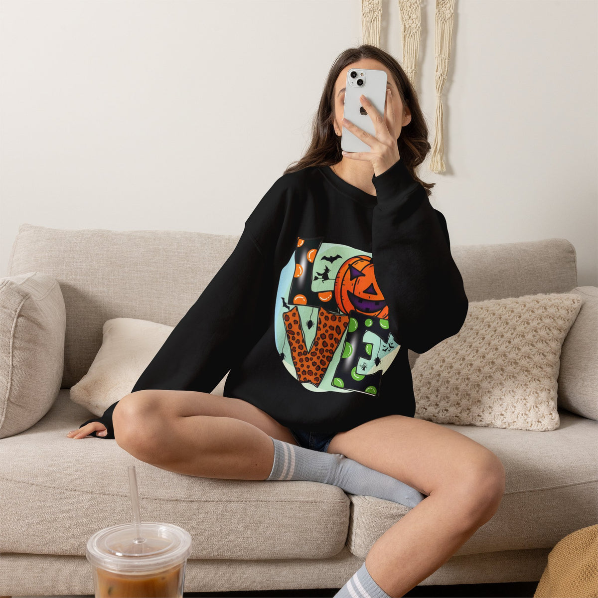 A woman sits on a beige couch taking a mirror selfie while wearing a black crewneck sweatshirt with a colorful graphic print of the word 'Halloween' in leopard print, orange, and green. She is wearing gray striped socks and has an iced coffee in the foreground.