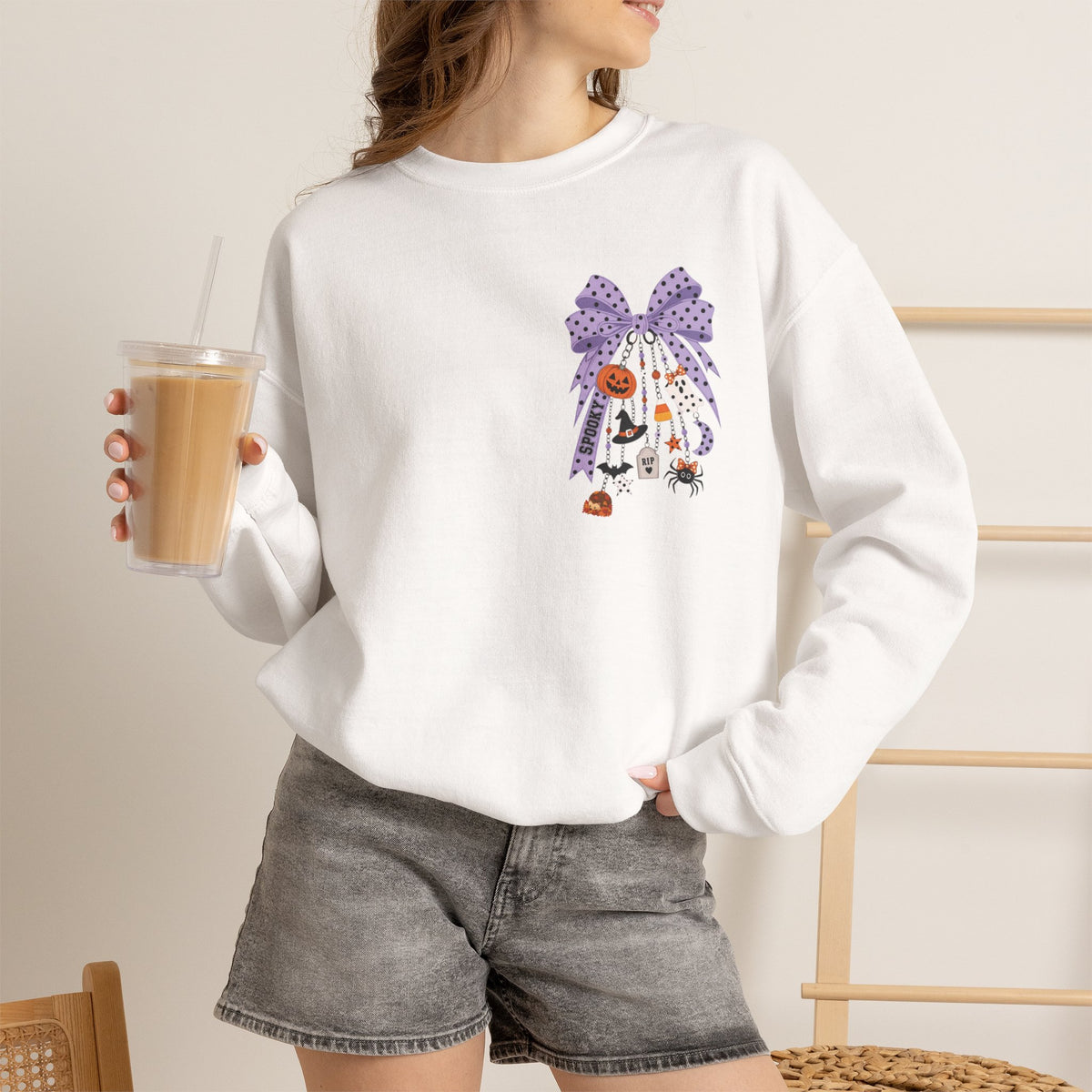 A woman wearing a white crewneck sweatshirt with a purple and orange Halloween-themed graphic print, paired with gray denim shorts and holding an iced coffee.