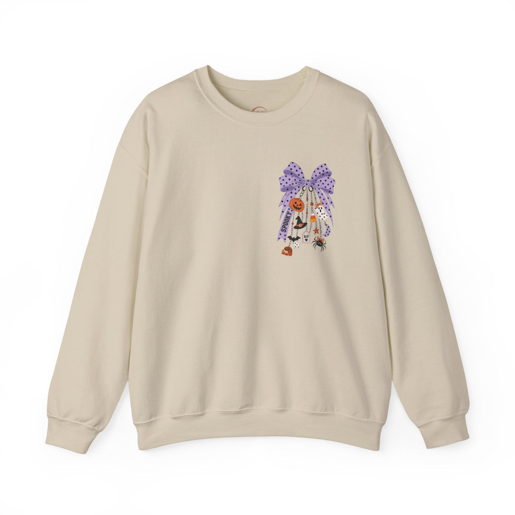 A beige crewneck sweatshirt featuring a whimsical embroidered design on the left chest of a purple patterned bow, a white cat, and small orange flowers, displayed against a clean white background.