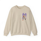 A beige crewneck sweatshirt featuring a whimsical embroidered design on the left chest of a purple patterned bow, a white cat, and small orange flowers, displayed against a clean white background.