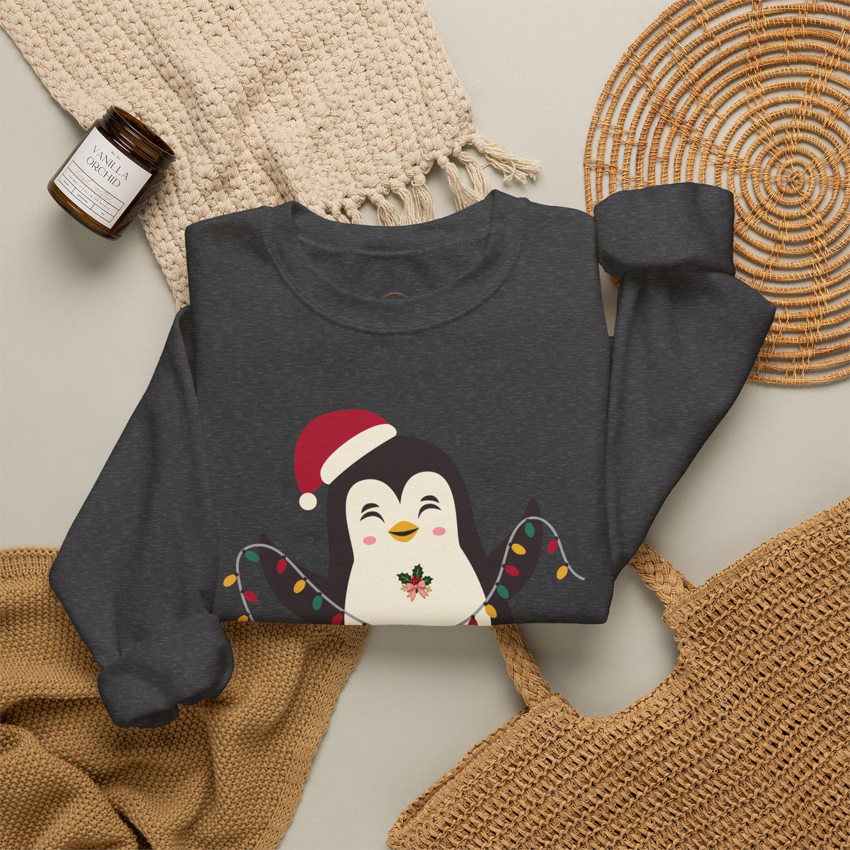 A dark gray crewneck sweatshirt featuring a cute cartoon penguin wearing a red Santa hat and holiday string lights, styled in a cozy flat-lay with a knit blanket, a woven placemat, and a small candle.