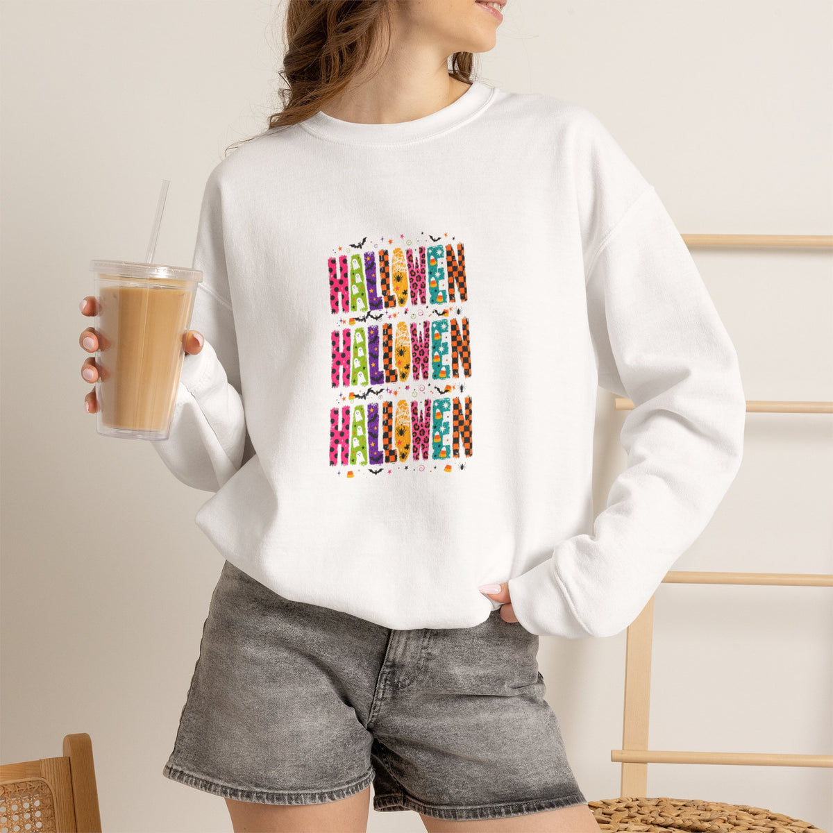A woman wearing a white crewneck sweatshirt with a colorful graphic print of the word 'HARSHAD' in stylized vertical lettering, paired with gray denim shorts and holding an iced coffee.