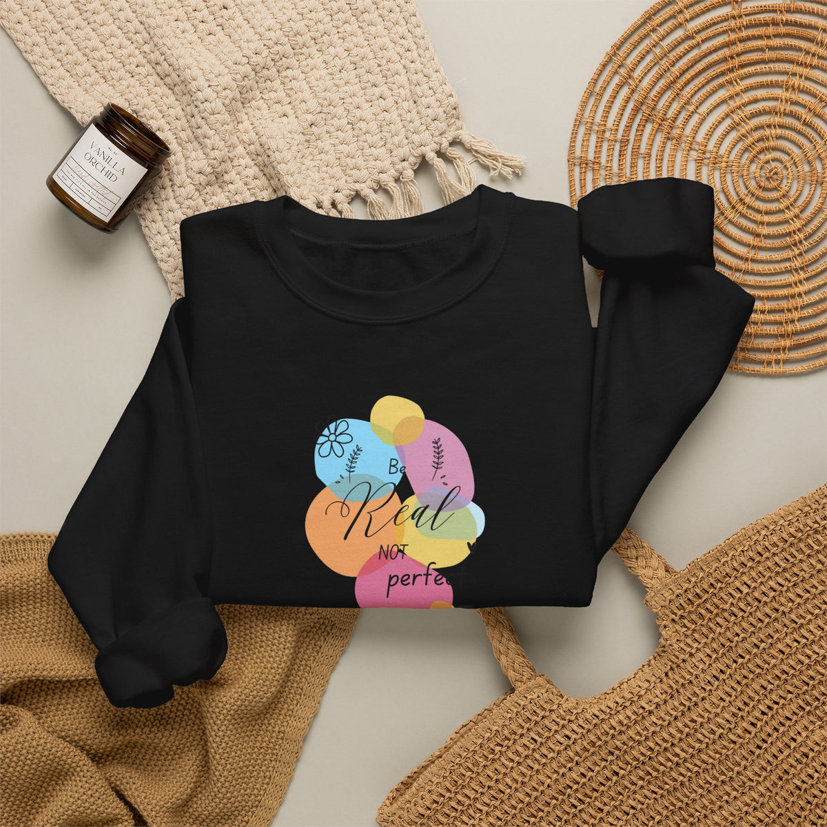 A black long-sleeve sweatshirt laid flat on a light beige surface, styled with a cream knit blanket, a woven placemat, a tan woven bag, and a small amber candle jar. The sweatshirt has a colorful pastel graphic of overlapping balloons with the words 'Be Kind' and 'NOT perfe' printed on them.