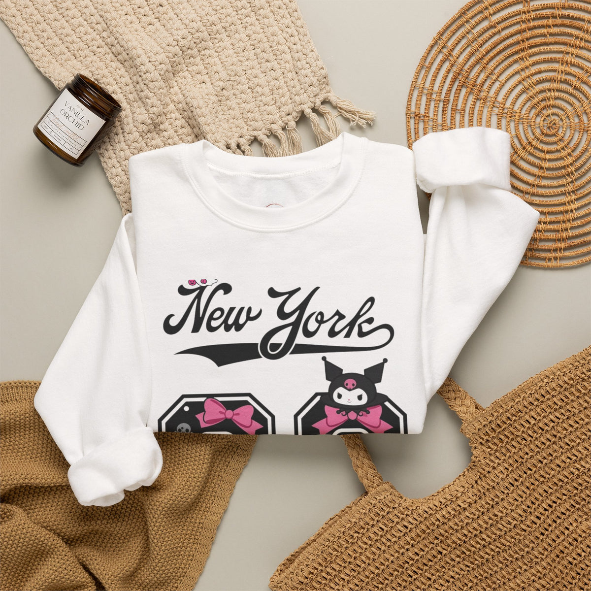 A folded white long-sleeve sweatshirt with a black 'New York' script graphic and cartoon character prints, styled in a flat lay with a woven blanket, a small candle jar, and a wicker tray on a light neutral background.