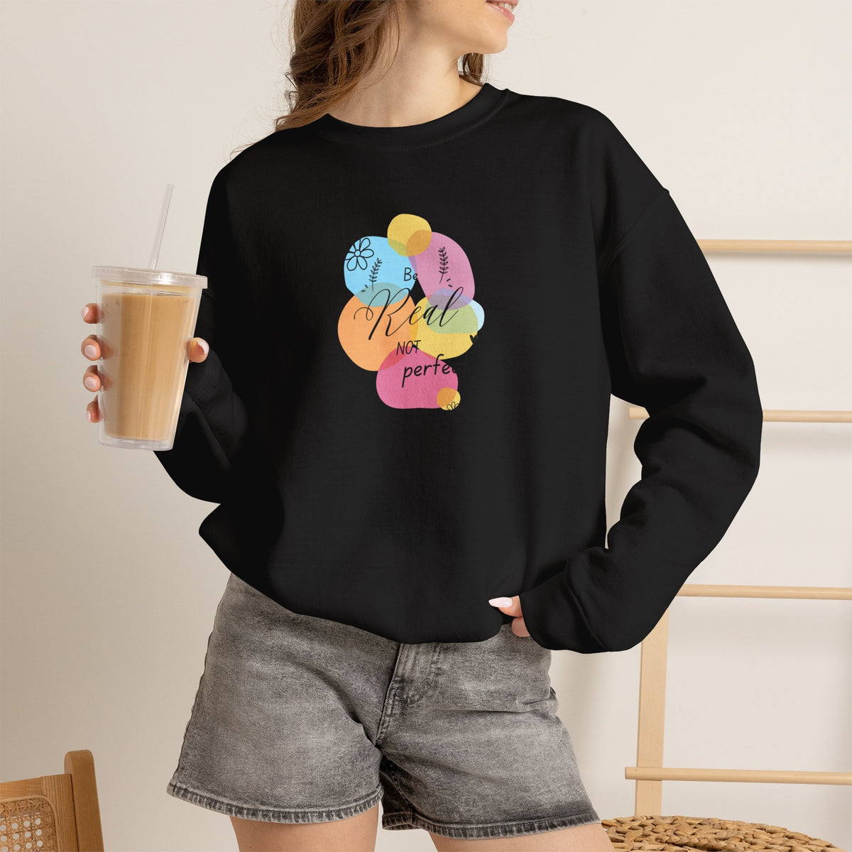 A woman wearing a black crewneck sweatshirt with a colorful graphic of baseball terms and the text 'Kub is not perfect' on the front, paired with gray denim shorts and holding an iced coffee.