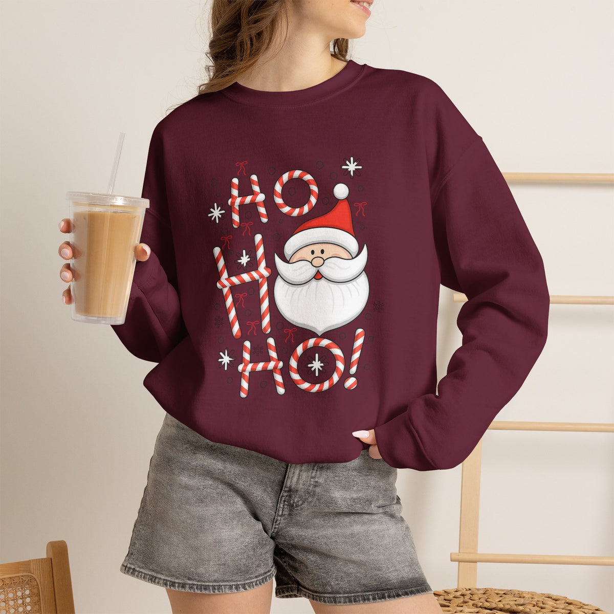 A woman wearing a maroon crewneck sweatshirt with a festive 'Ho Ho Ho' Santa graphic, paired with gray denim shorts and holding an iced coffee.
