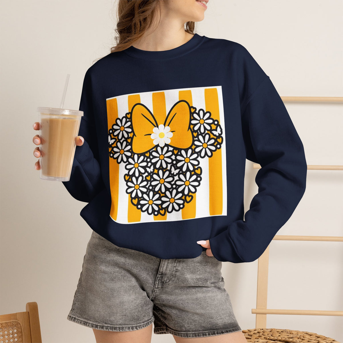 A woman wearing a navy blue sweatshirt with a graphic print of a yellow bow, black and white daisies, and yellow-and-white vertical stripes. She is holding an iced coffee and wearing gray denim shorts in a brightly lit indoor setting.