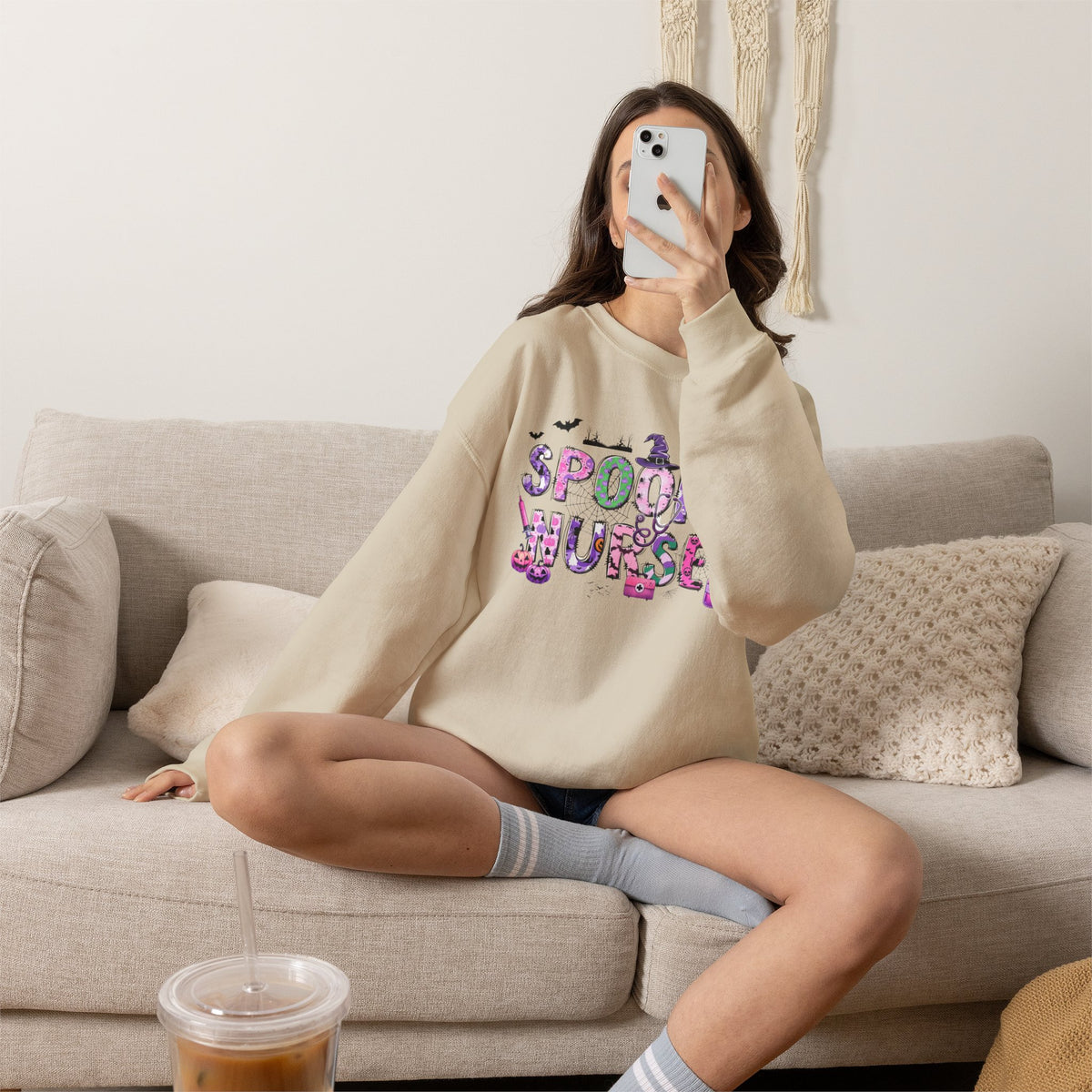 A woman takes a mirror selfie while wearing a beige crewneck sweatshirt with a colorful 'SPOOKY CURSE' graphic on the front, sitting on a light beige couch.