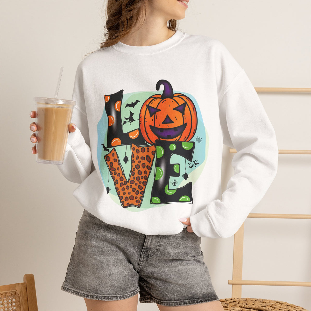 A woman wearing a white crewneck sweatshirt with a colorful Halloween-themed 'LOVE' graphic featuring a jack-o'-lantern and bats, paired with gray denim shorts and holding an iced coffee.