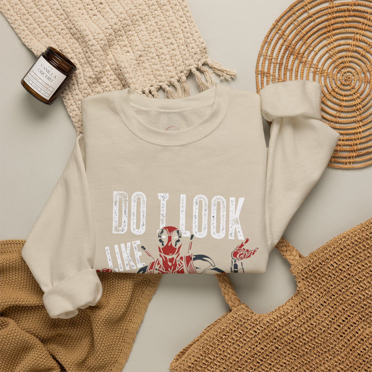 A cream-colored crewneck sweatshirt featuring a distressed graphic that reads 'DO I LOOK LIKE' above a red and black cartoon character, styled in a flat lay with a woven blanket, a small candle, and a wicker tray.
