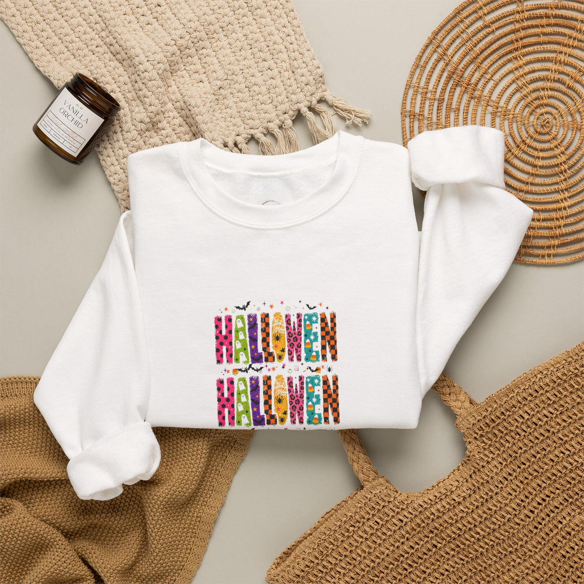 A white long-sleeve crewneck sweatshirt with a colorful 'HALLOWEEN' graphic made of patterned letters, styled in a flat lay with a knit blanket, a woven mat, and a candle.