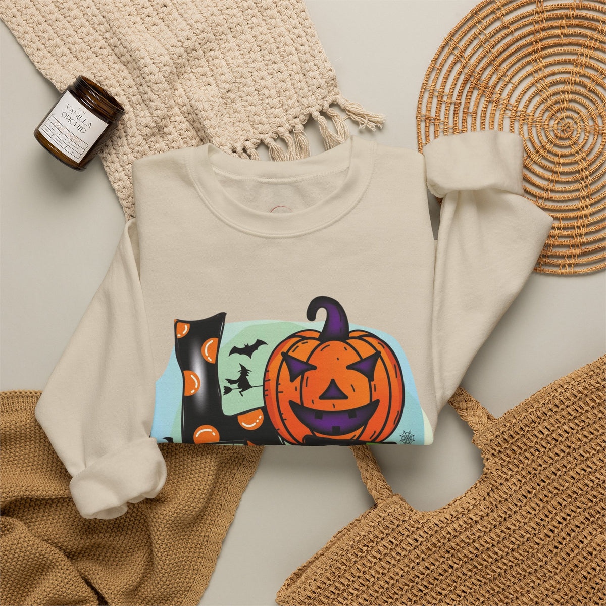 A cream-colored crewneck sweatshirt featuring a colorful Halloween graphic with a jack-o'-lantern, witch hat, bats, and pumpkins, styled in a flat lay with a knit blanket, woven placemat, and a candle.