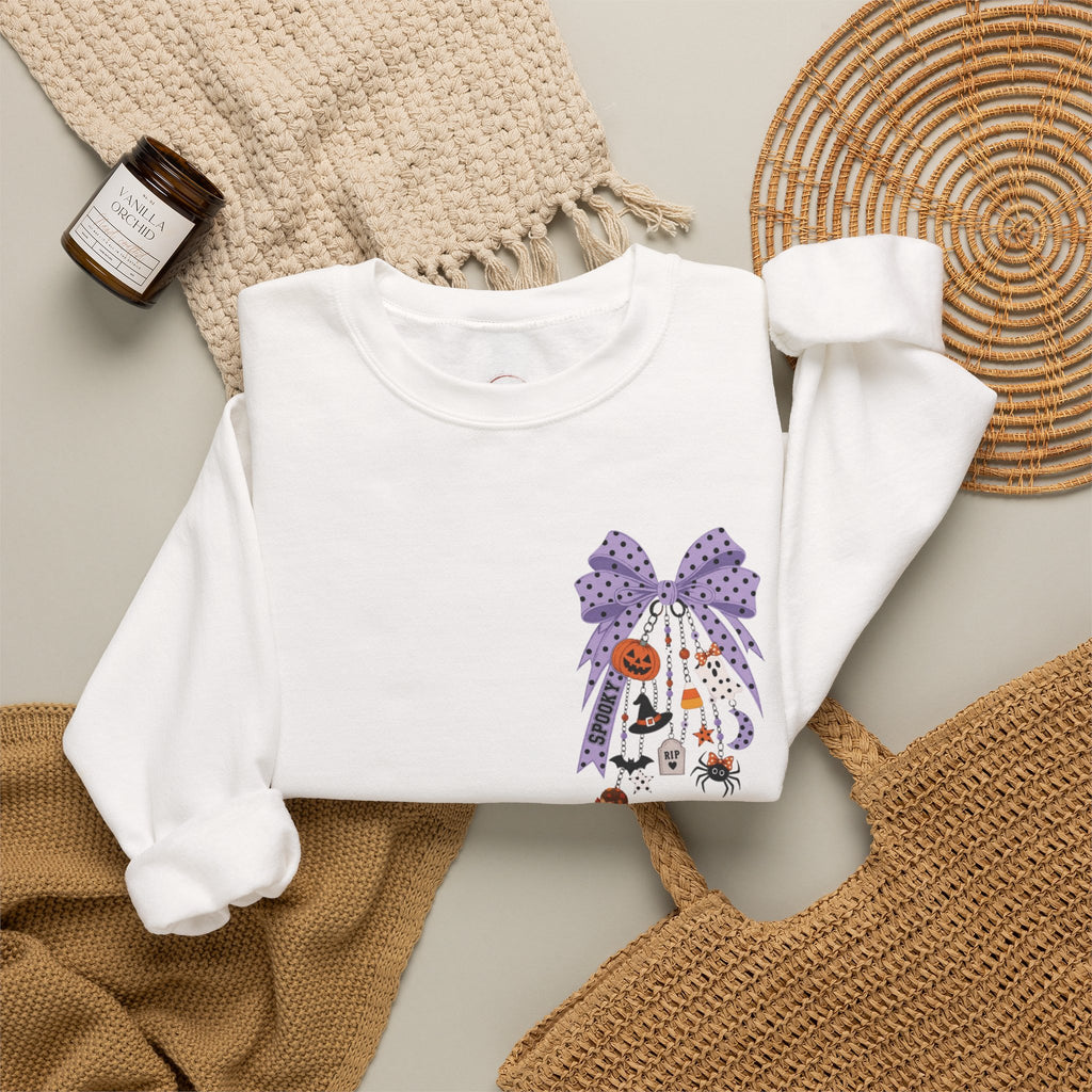A white long-sleeve crewneck sweatshirt with a purple Halloween-themed graphic print, styled in a flat lay with a woven blanket, a candle, a wicker tray, and a tan tote bag.
