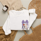 A white long-sleeve crewneck sweatshirt with a purple Halloween-themed graphic print, styled in a flat lay with a woven blanket, a candle, a wicker tray, and a tan tote bag.