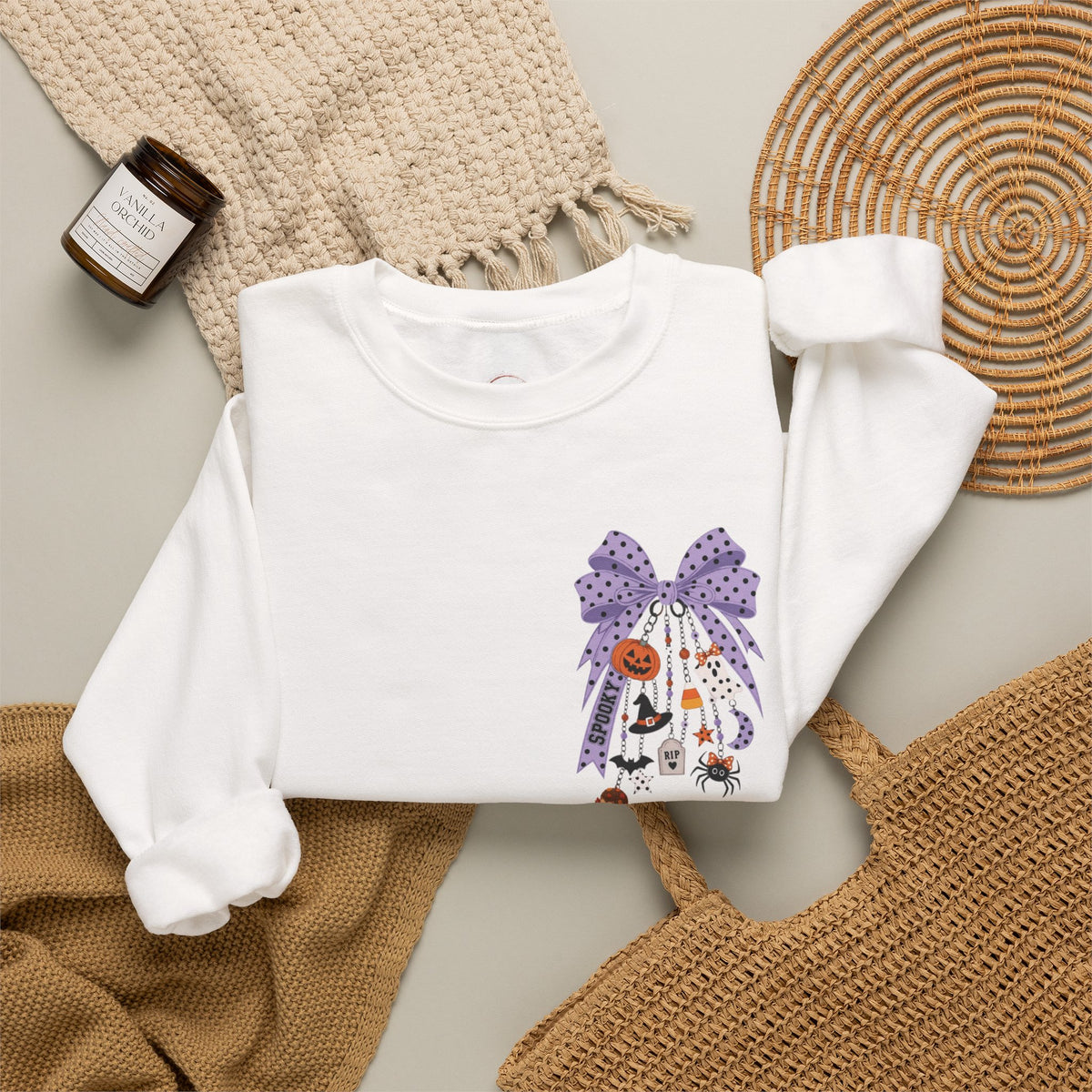 A white long-sleeve crewneck sweatshirt with a purple Halloween-themed graphic print, styled in a flat lay with a woven blanket, a candle, a wicker tray, and a tan tote bag.